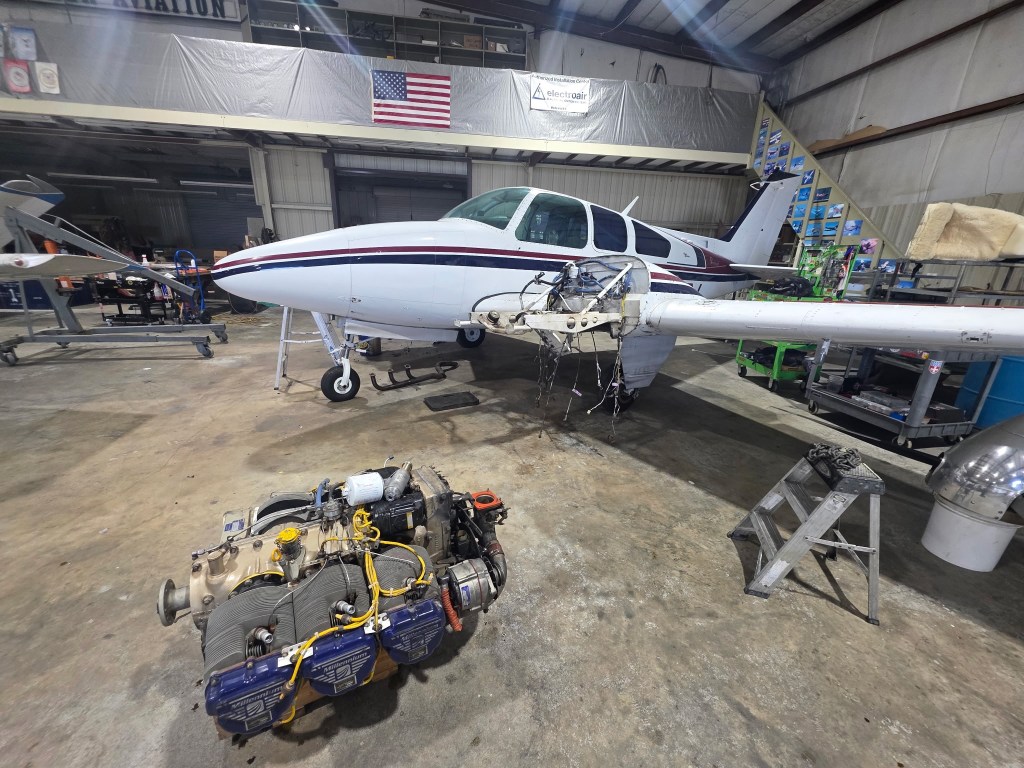 View of a Beech Baron in a hangar, with one engine removed and sitting on the floor in front of it
